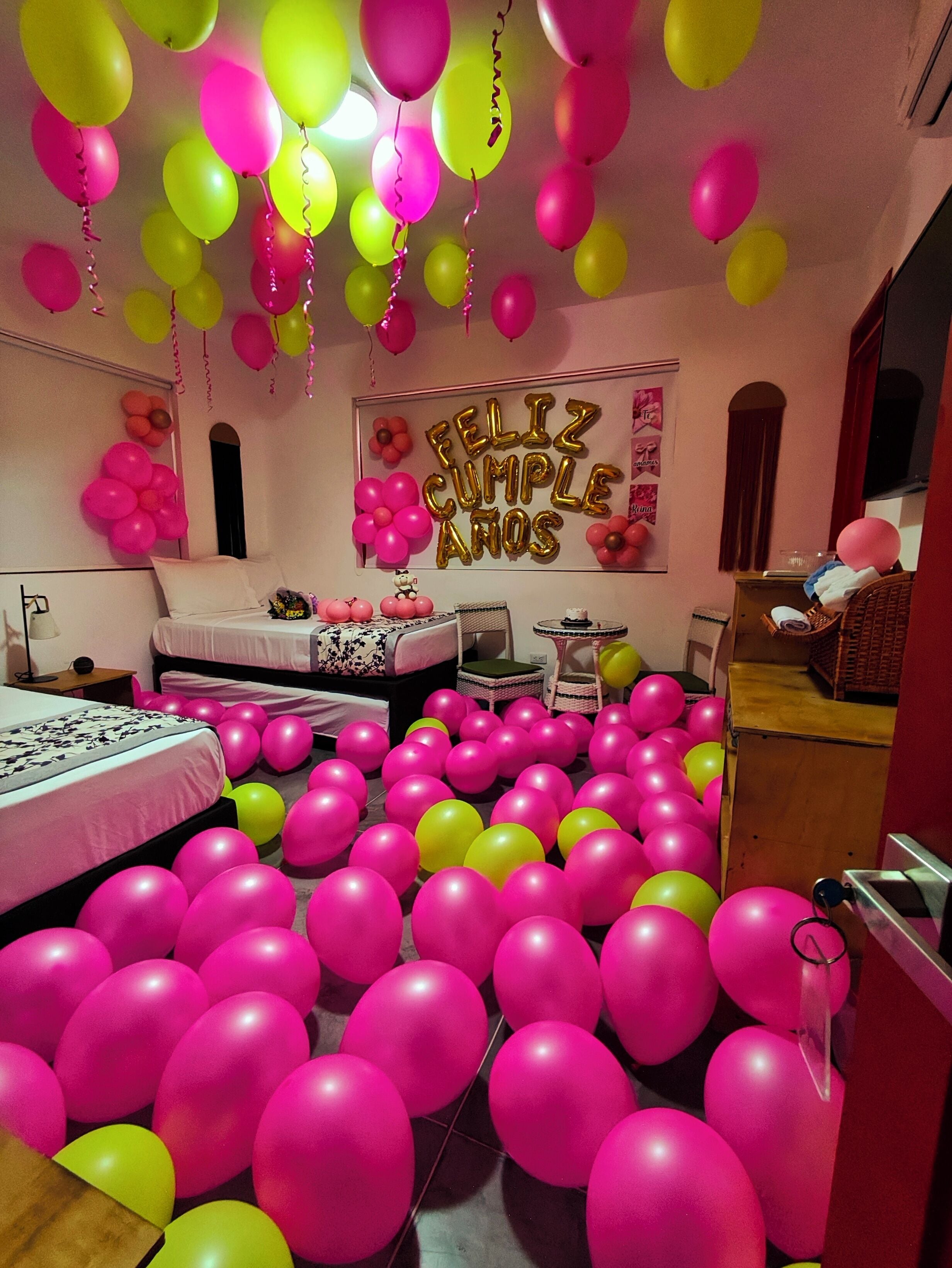 Een kamer met roze en gele ballonnen, een bed, een kleine tafel en een wanddecoratie met de tekst 'Feliz Cumple Años'.