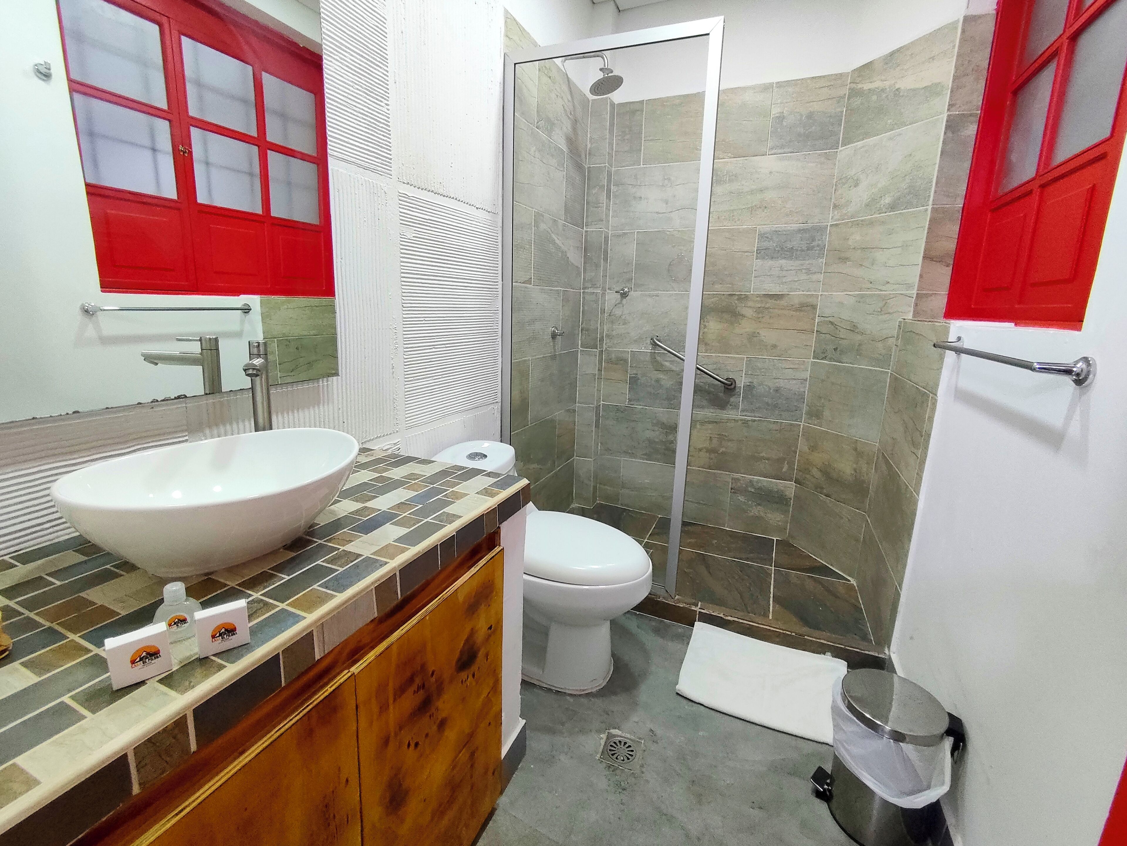 A bathroom with a large white sink, a toilet, a shower with stone tiles, and a red-framed mirror.