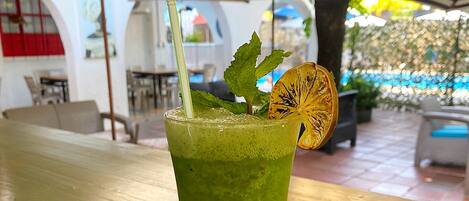 Una bebida verde con pajita y una rodaja de fruta seca como adorno, sobre una mesa de madera en un área de estar al aire libre.