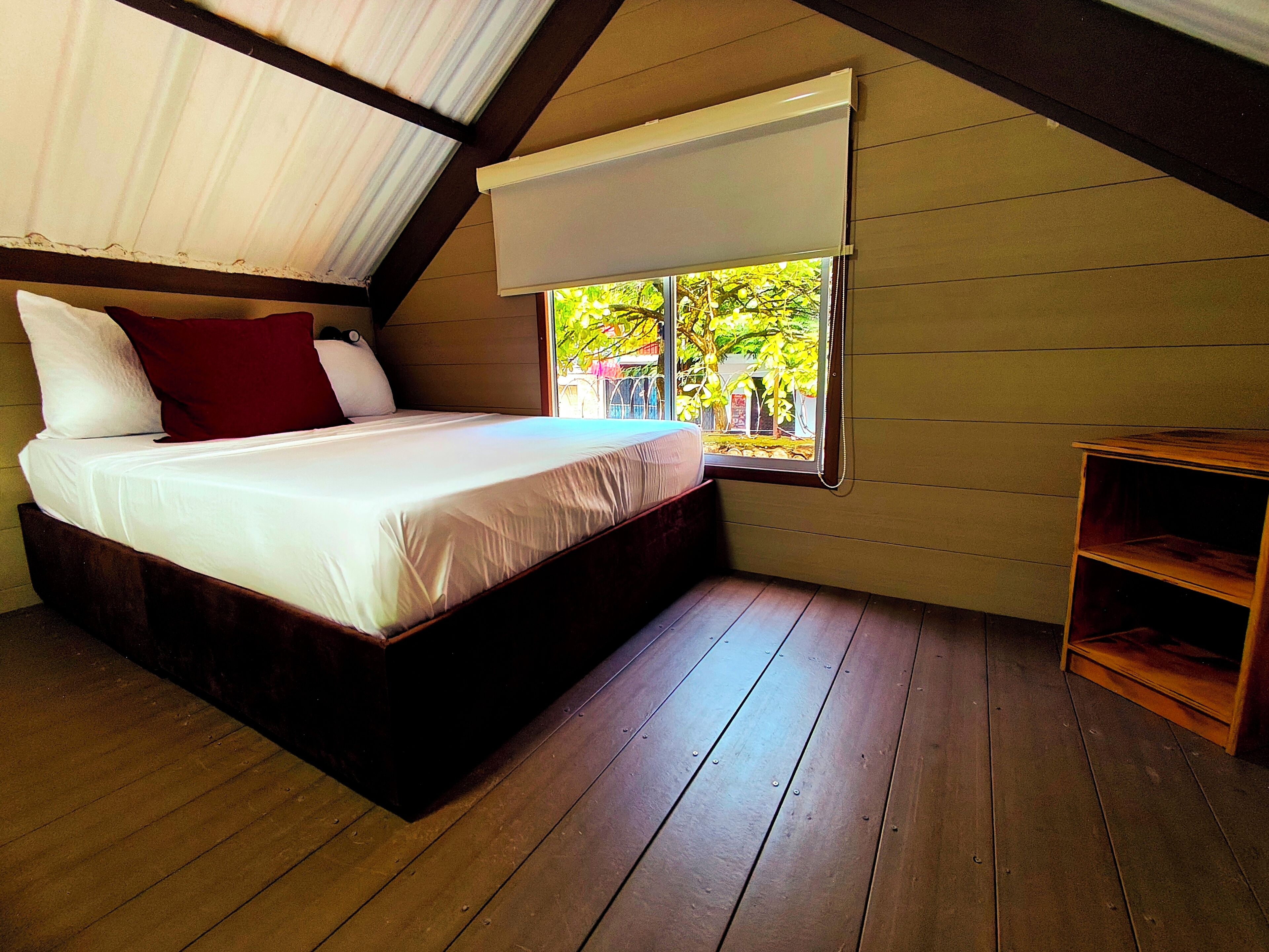 Un dormitorio con piso de madera, una cama con ropa blanca, un almohadón rojo y una ventana con vistas al verdor.