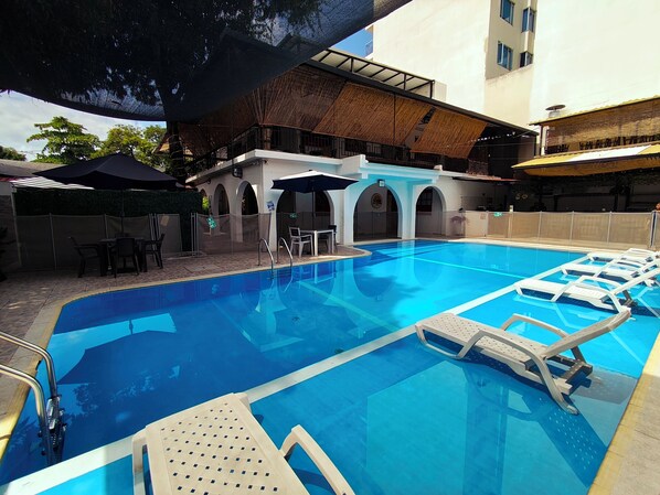 Una piscina con sillones de descanso, una zona de estar techada y un patio cercado.