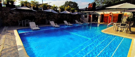 Una piscina con agua azul cristalina, rodeada de sillas de descanso y sombrillas, con un muro de piedra y una torre de salvavidas al fondo.