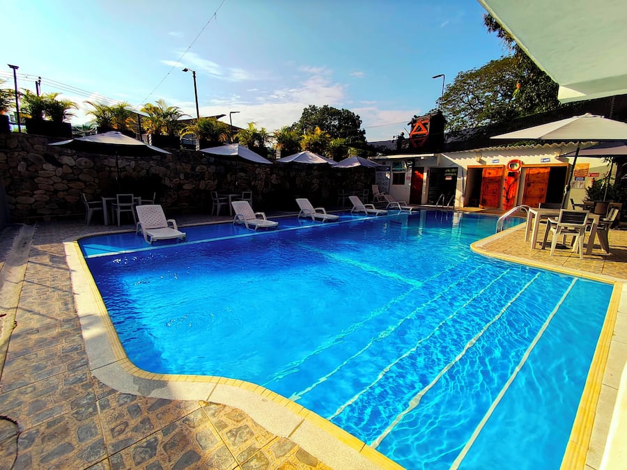 A swimming pool with clear blue water, surrounded by lounge chairs and umbrellas, with a stone wall and a lifeguard tower in the background.