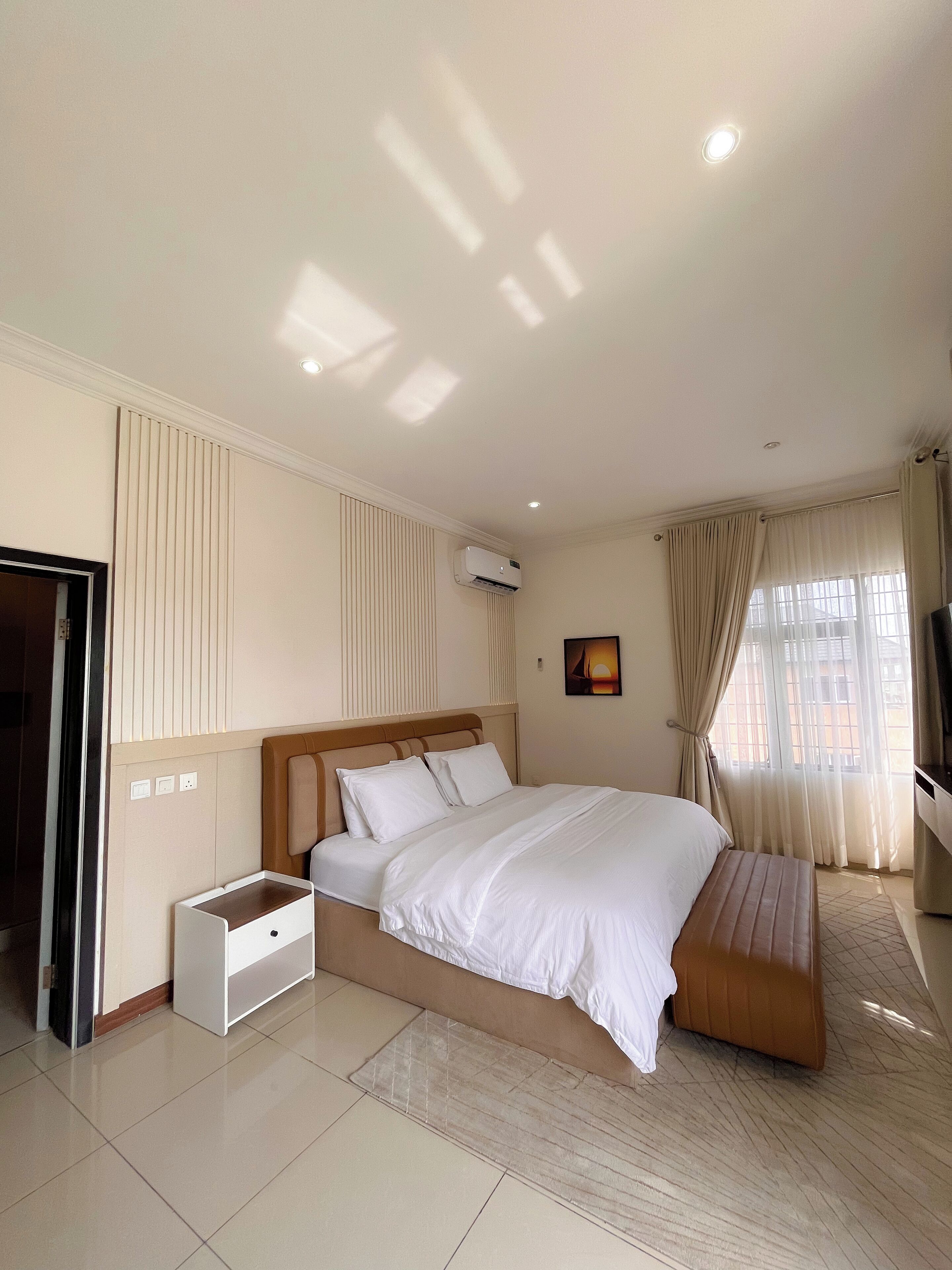 A hotel room with a large bed, a bedside table, an air conditioning unit, a TV, and a window with curtains.