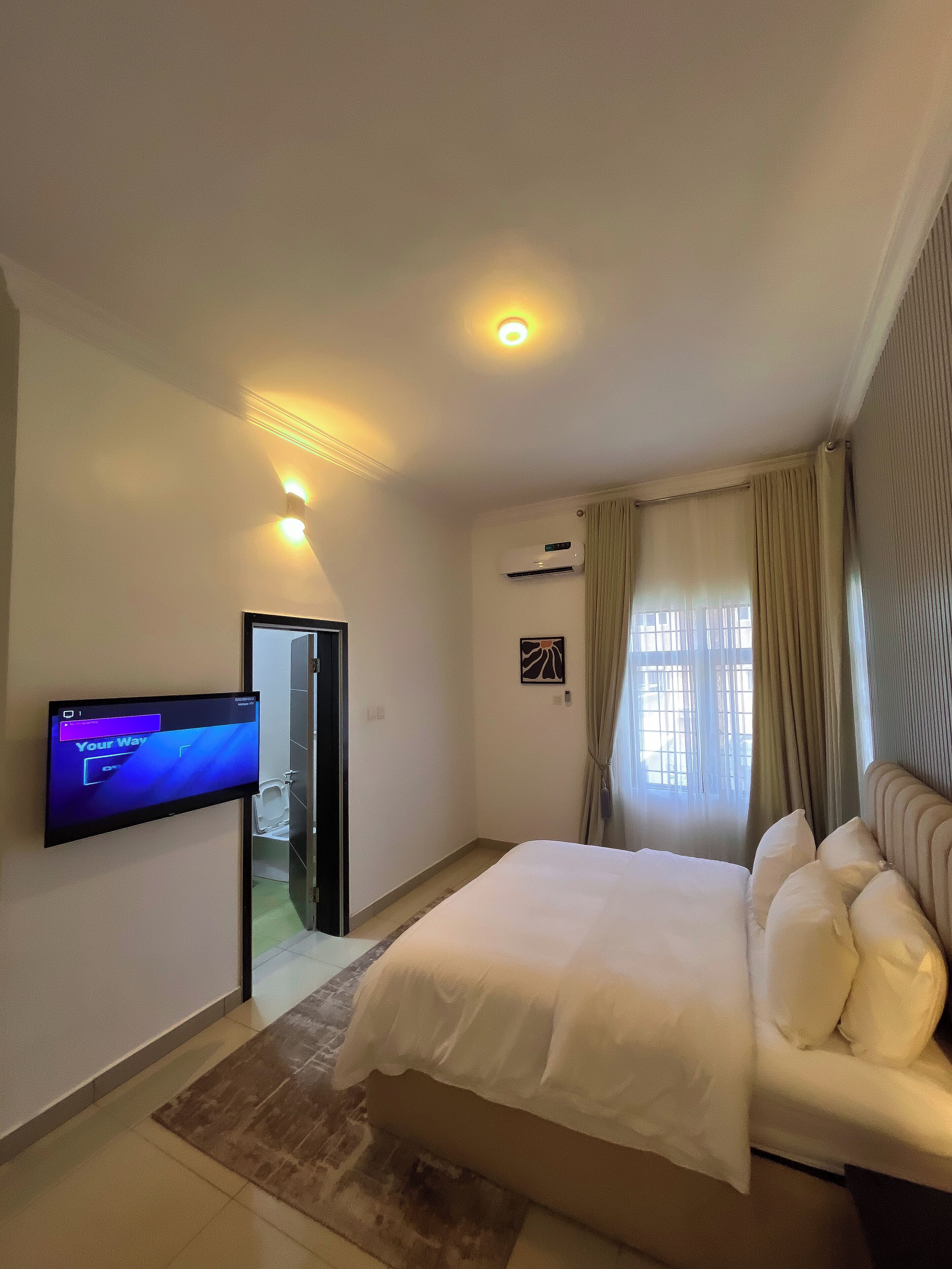 A hotel room with a bed, a flat-screen TV mounted on the wall, an air conditioning unit, and a window with curtains.