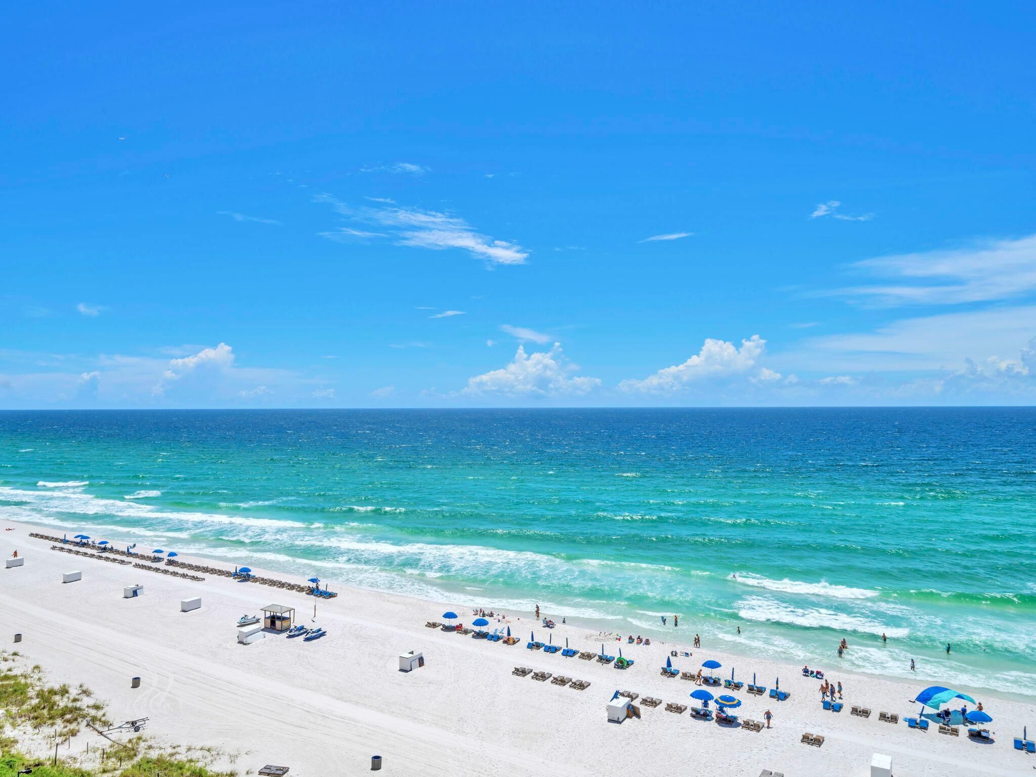 A beach with white sand, clear blue water, and a few umbrellas and lounge chairs.