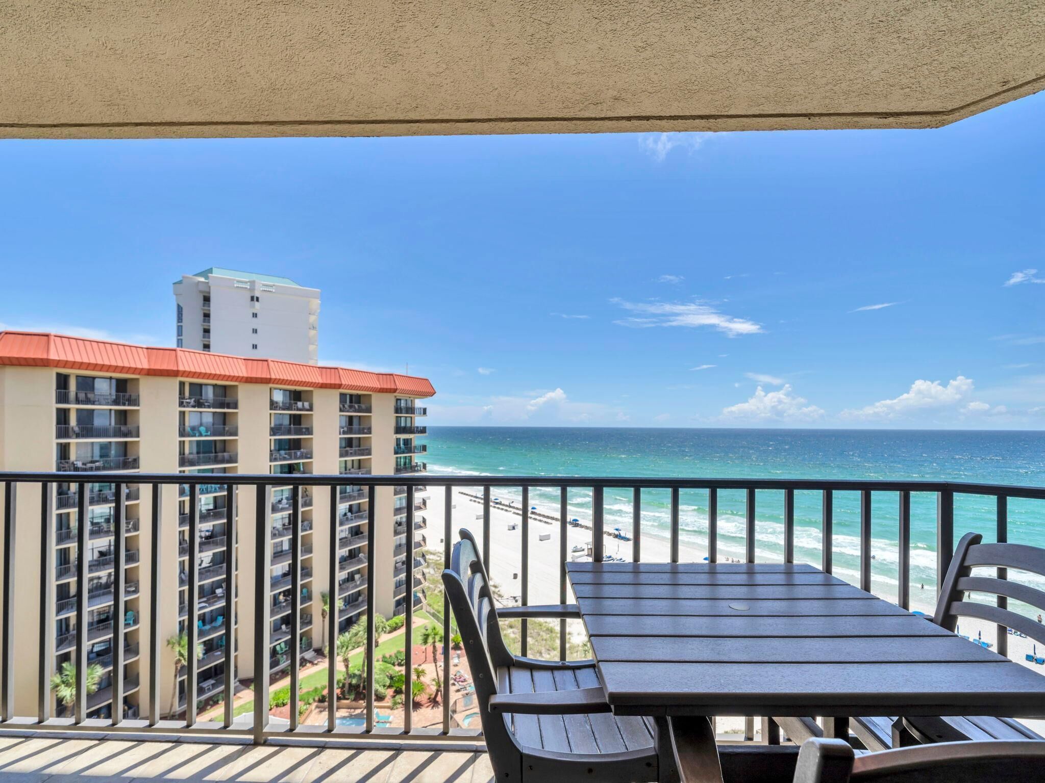 Un balcon avec vue sur la plage et l’océan, une table et, au loin, un immeuble de grande hauteur.