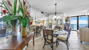 A spacious dining area with a glass table and wicker chairs, a wooden console table with plants, and a view of the ocean through large windows. - Summerhouse 1012c (Panama City Beach)