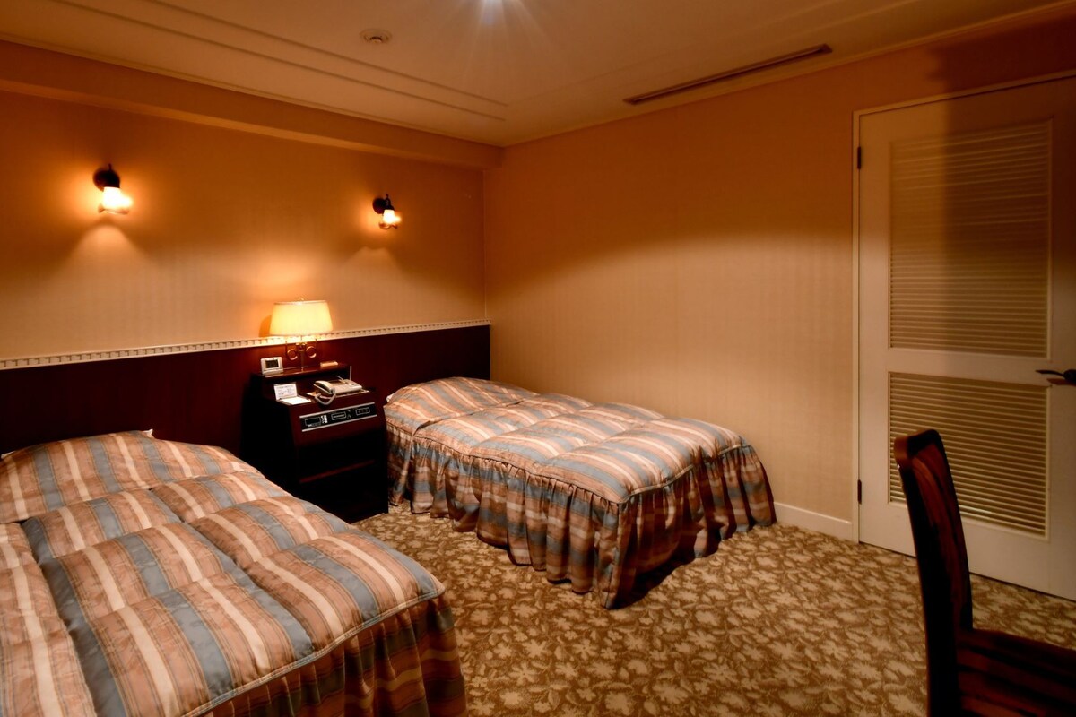 Habitación de hotel con dos camas, una mesita de noche, una lámpara y una puerta con ventanal.