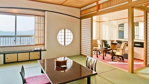 Una habitación tradicional japonesa con suelo de tatami, una mesa baja con cojín y puertas correderas que dan a un balcón con vistas a la montaña.