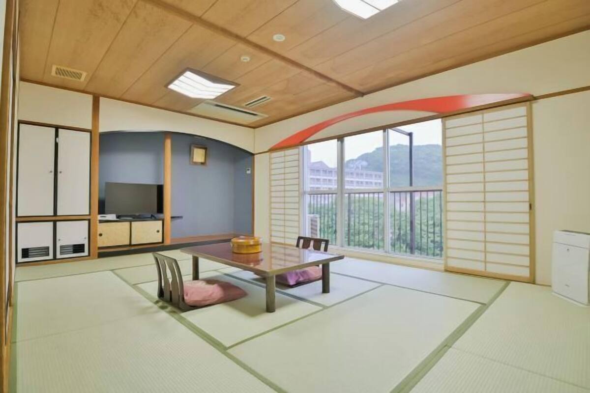 A traditional Japanese room with tatami flooring, a low table, a TV, sliding doors, and a view of buildings.
