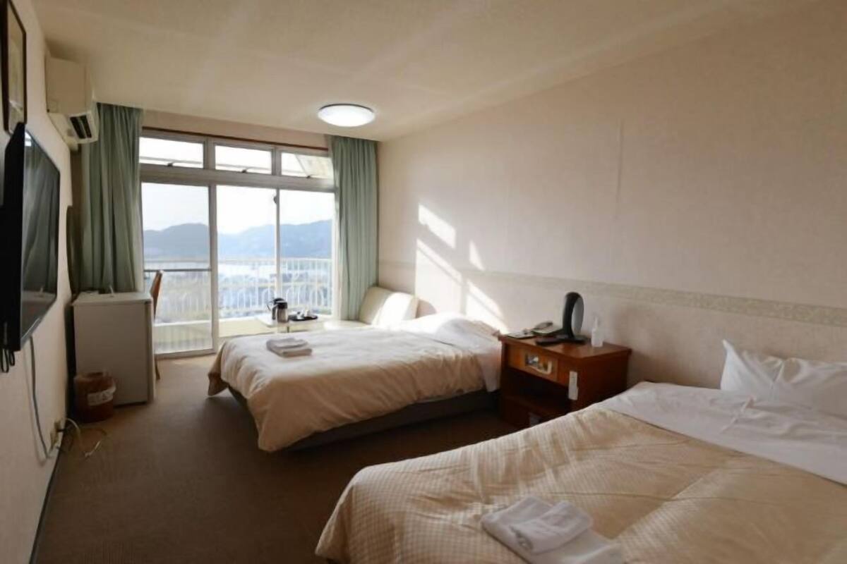 A hotel room with two beds, a small desk, a chair, a window with a view, and a ceiling light.