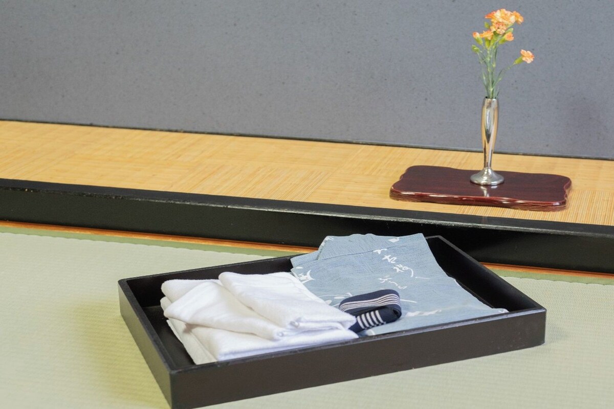 Zona de recepción de hotel con una mesita negra que sostiene toallas dobladas y un objeto a rayas, un florero con flores sobre una superficie de madera, y un objeto rojo oscuro sobre un soporte.