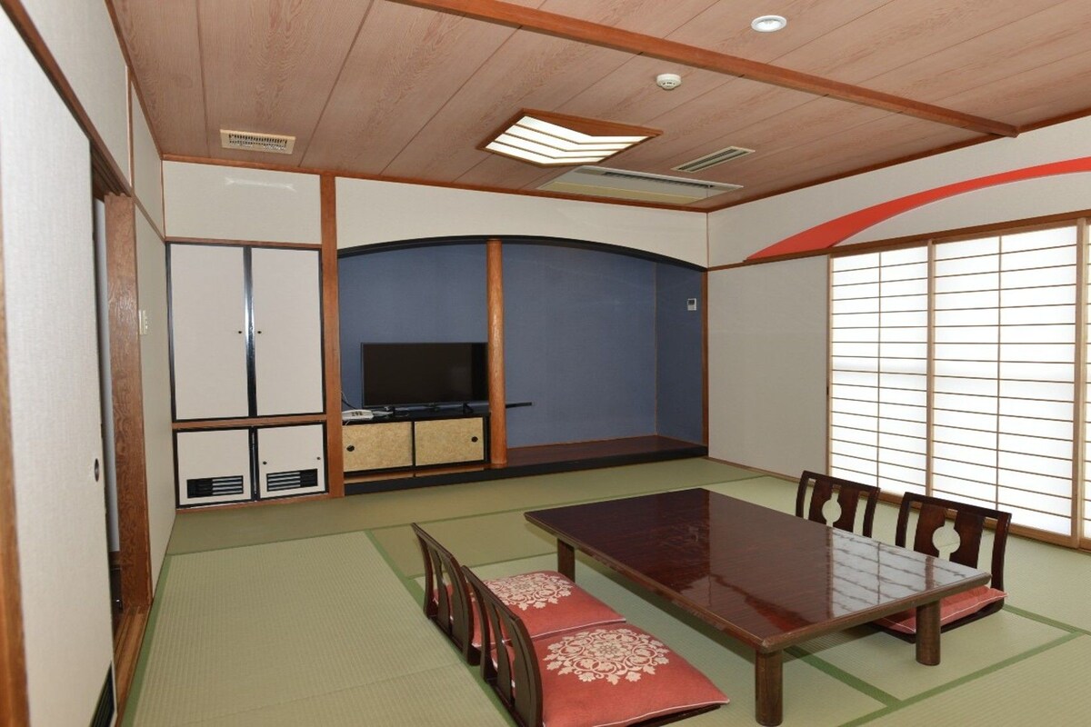 Una habitación de estilo japonés tradicional con suelo de tatami, una mesa baja con sillas, un televisor y puertas correderas.