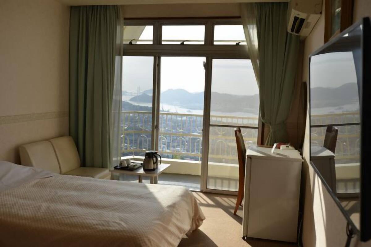 A hotel room with a bed, a desk, a chair, and a TV. There is a large window with a view of a bridge and mountains.