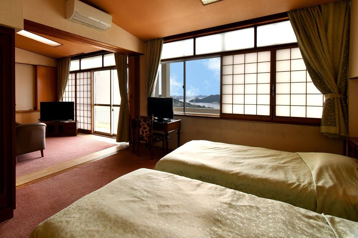 A hotel room with two beds, a TV, a desk, and large windows with curtains.