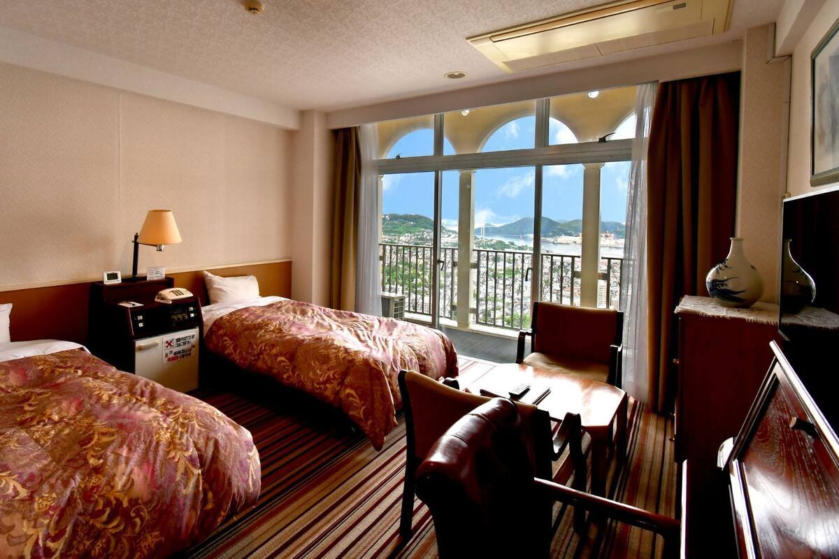 A hotel room with two beds, a desk, a chair, a nightstand, and a large window with a view.