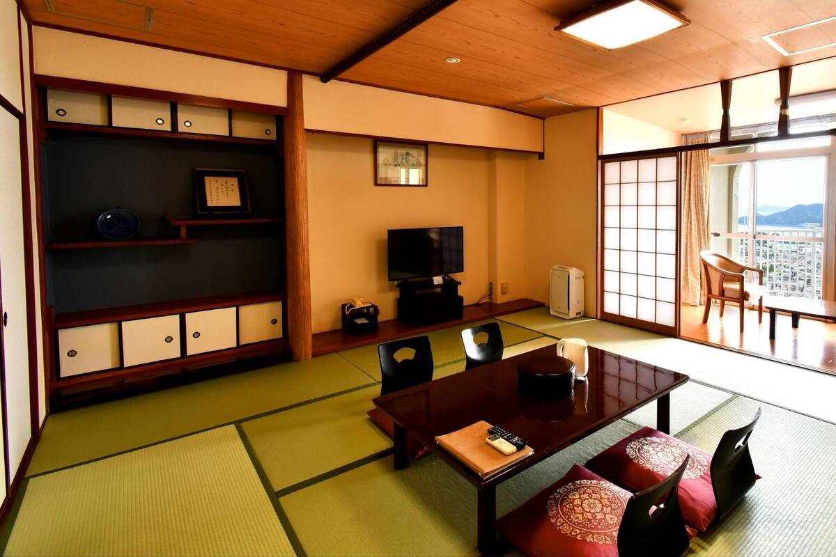 A traditional Japanese room with tatami flooring, a low wooden table, a television, and a view of a cityscape through large shoji doors.