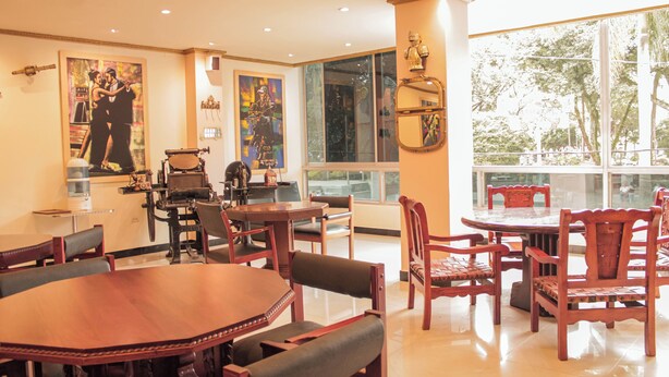 A dining area with wooden tables and chairs, large windows with a view of trees, and artwork on the walls. - Hotel Plaza Garibaldi (Caldas)