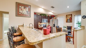 A modern kitchen with granite countertops, dark wooden cabinets, a stainless steel refrigerator, and a microwave. There is a dining area with a table and chairs.