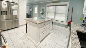 Una cocina moderna con armarios blancos, un refrigerador de acero inoxidable, encimera de granito y ventana con persianas.