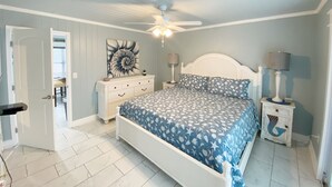 Un dormitorio con cama, ventilador de techo, mesita de noche, cómoda y una obra de arte en la pared.