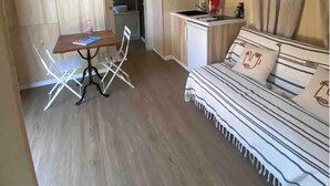 A compact room with a wooden ceiling, a dining table with chairs, a sofa with striped bedding, and a kitchenette area.