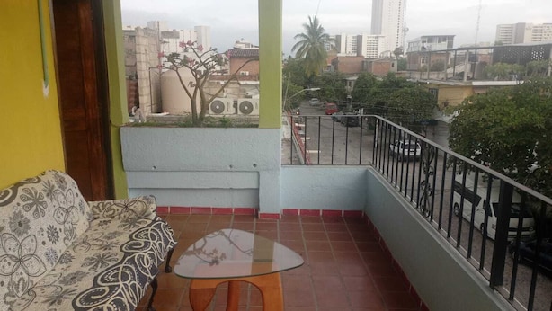A balcony with a sofa and a small table, overlooking a cityscape with buildings and a palm tree.