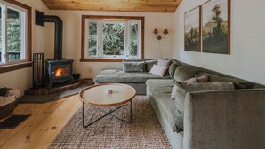 A cozy living room with a fireplace, a green sofa, a round wooden coffee table, and a large window with a view of trees.