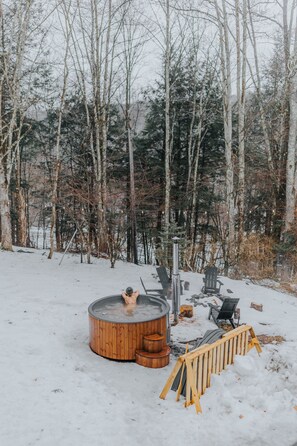 A wooden hot tub surrounded by snow in a forested area.