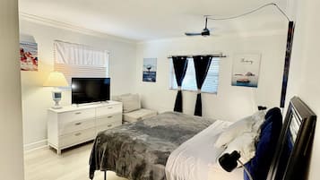 A bedroom with a bed, bedside table, lamp, TV, and a window with curtains.