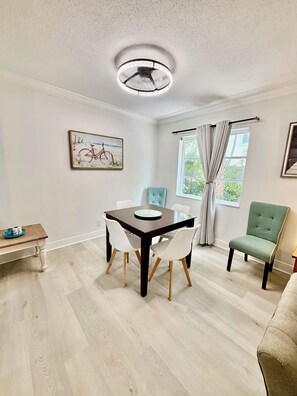 A dining area with a table, chairs, and a bench. There is a painting on the wall and a window with curtains.