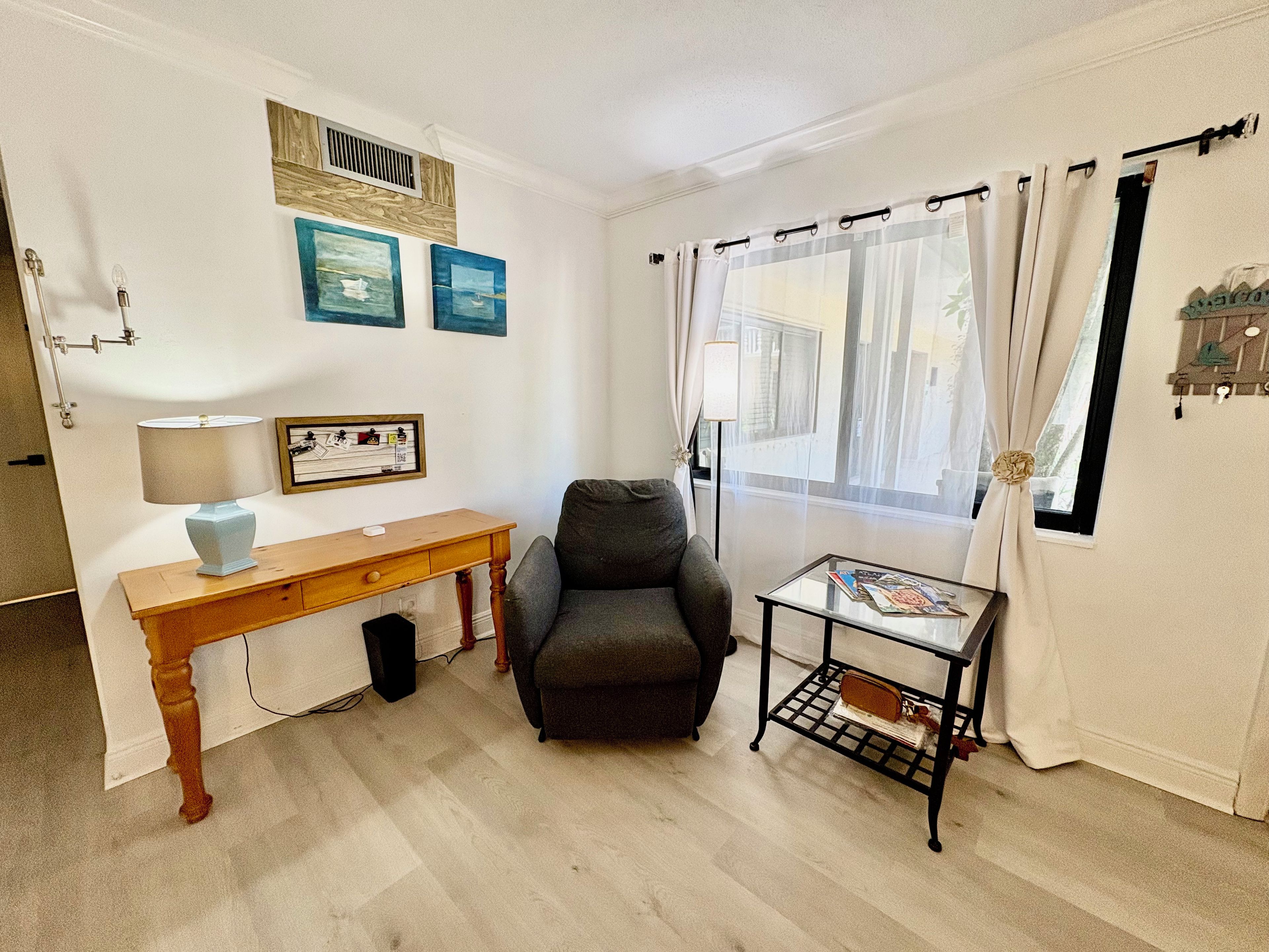 A room with a wooden desk, a grey armchair, a small table with magazines, and a window with curtains.
