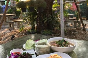 A table with a meal consisting of rice, a bowl of soup, and a plate of food, set outdoors with a tree and plants in the background.