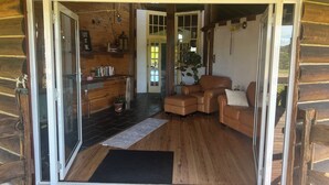 A cozy cabin interior with a wooden floor, a leather sofa, a dining area with a table and chairs, and a large window offering a view of the outdoors. - Log Cabin OFF GRID, fire place, balconies/library/ Rain forrest, walk, swim.  (Elands)