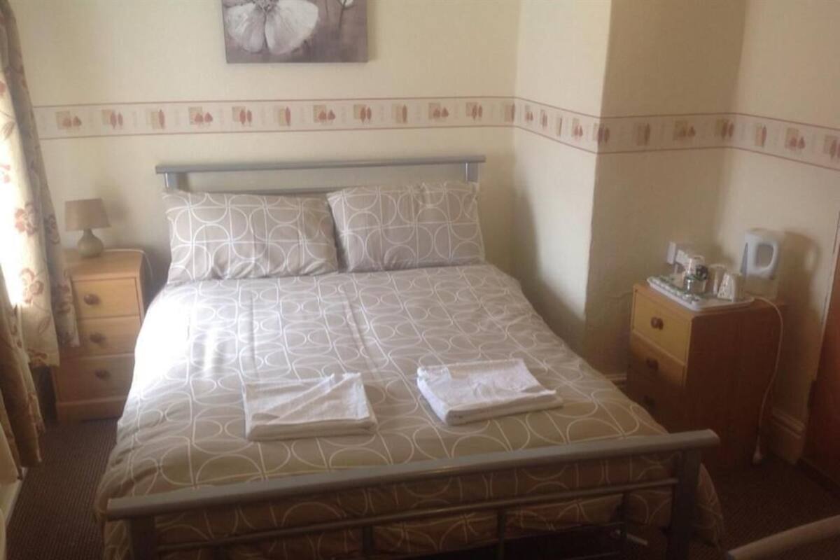 Een net opgemaakt bed met decoratieve beddengoed, een nachtkastje met een lamp, en een tafeltje met een koffiezetapparaat.
