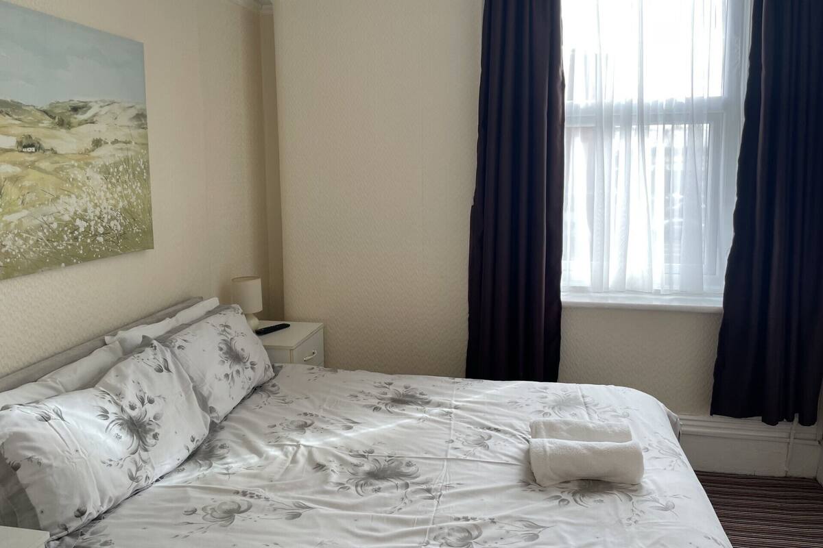 Een opgemaakt bed met bloemenbedrukking, een nachtkastje met een lamp, en een schilderij aan de muur.