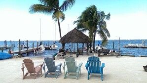 A beachside area with a thatched-roof hut, several wooden chairs, and a view of the sea with boats. - Bungalow w/ FREE Kayaks, Sailboat, Bicycle Use (Key Largo)