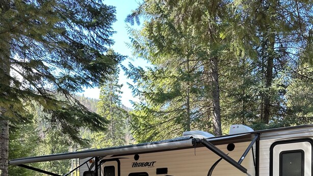 A campsite with a beige RV named "HIDEOUT" parked on a checkered mat. Two red and white folding chairs are set up on the mat. - Back Country Getaway. Complete 30/50 RV Site. (Kingston)