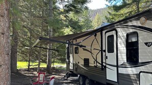 A recreational vehicle parked in a forested area with a checkered mat and red chairs. - Back Country Getaway. Complete 30/50 RV Site. (Kingston)