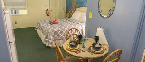 A compact room with a bed, a dining table with chairs, a mirror, and a wall painting.