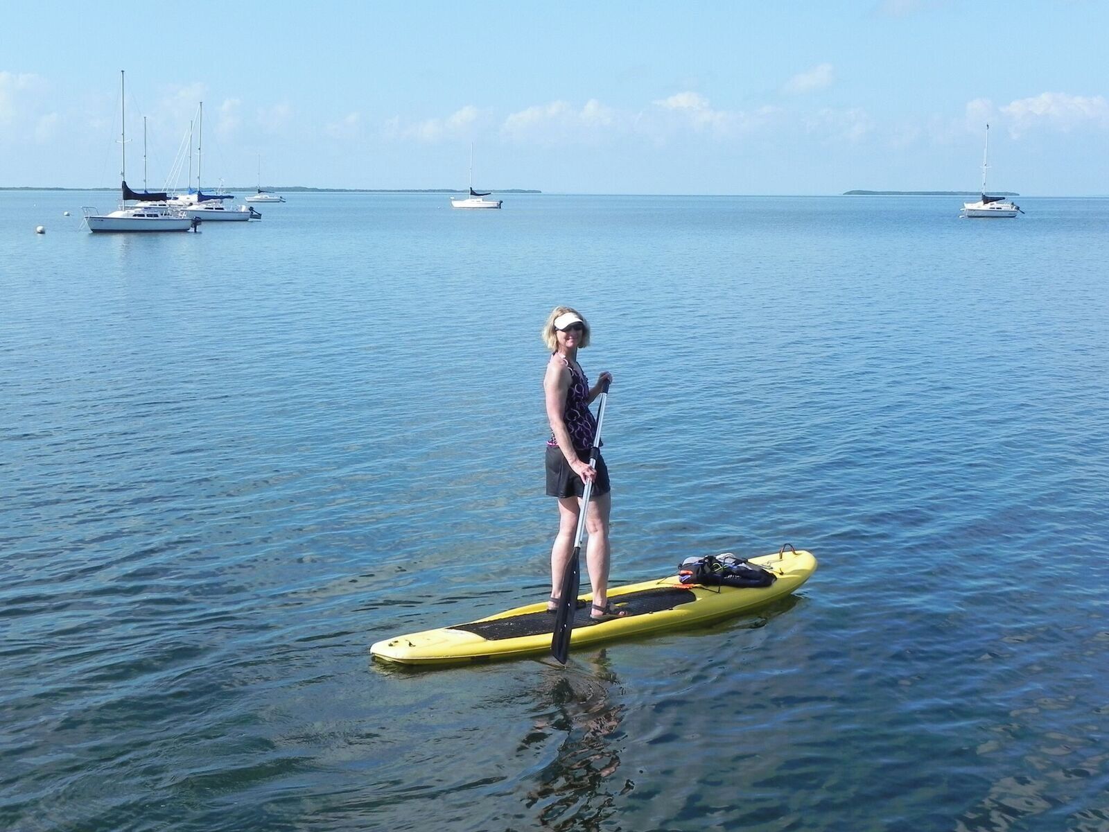 A person standing on a paddleboard in calm water with several boats in the background.