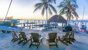 A beachside area with wooden chairs arranged in a semi-circle, a thatched-roof structure, and boats docked at a pier.