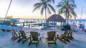 A beachside area with wooden chairs arranged in a semi-circle, a thatched-roof structure, and boats docked at a pier. - Bayside view Cottage , Free kayaks and sailboat (Key Largo)