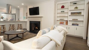 Un salón con chimenea, un sofá, una mesa de centro y un televisor montado sobre la chimenea. Se observan libros y objetos decorativos en estanterías.