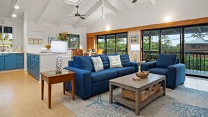 A modern living room with a blue sofa set, a wooden coffee table, and a glass-top table. There are ceiling fans, a skylight, and large windows offering a view of the outdoors. - Haena Hale Kauai Beach House (Hanalei)