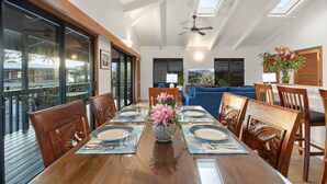 A dining area with a wooden table set for six, surrounded by chairs, with a view of the outdoors through large windows. - Haena Hale Kauai Beach House (Hanalei)