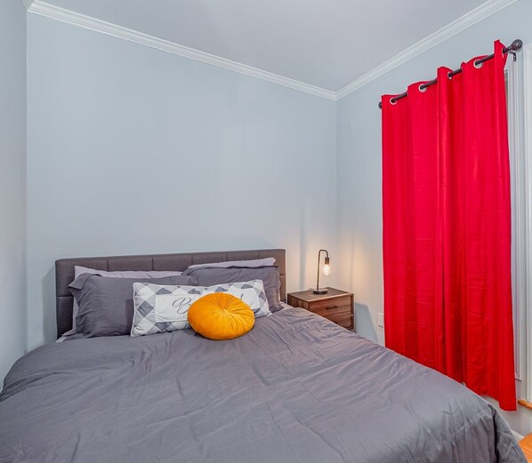 A bedroom with a bed, pillows, a nightstand, a lamp, and a red curtain.