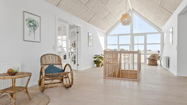 A spacious room with a high ceiling, large windows, a wicker chair, a small table with a bowl of fruit, and a view of the outdoors.