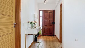 Un passadís amb sòl de fusta, una taula consola blanca amb un ram de flors, una porta de fusta i un interruptor a la paret.